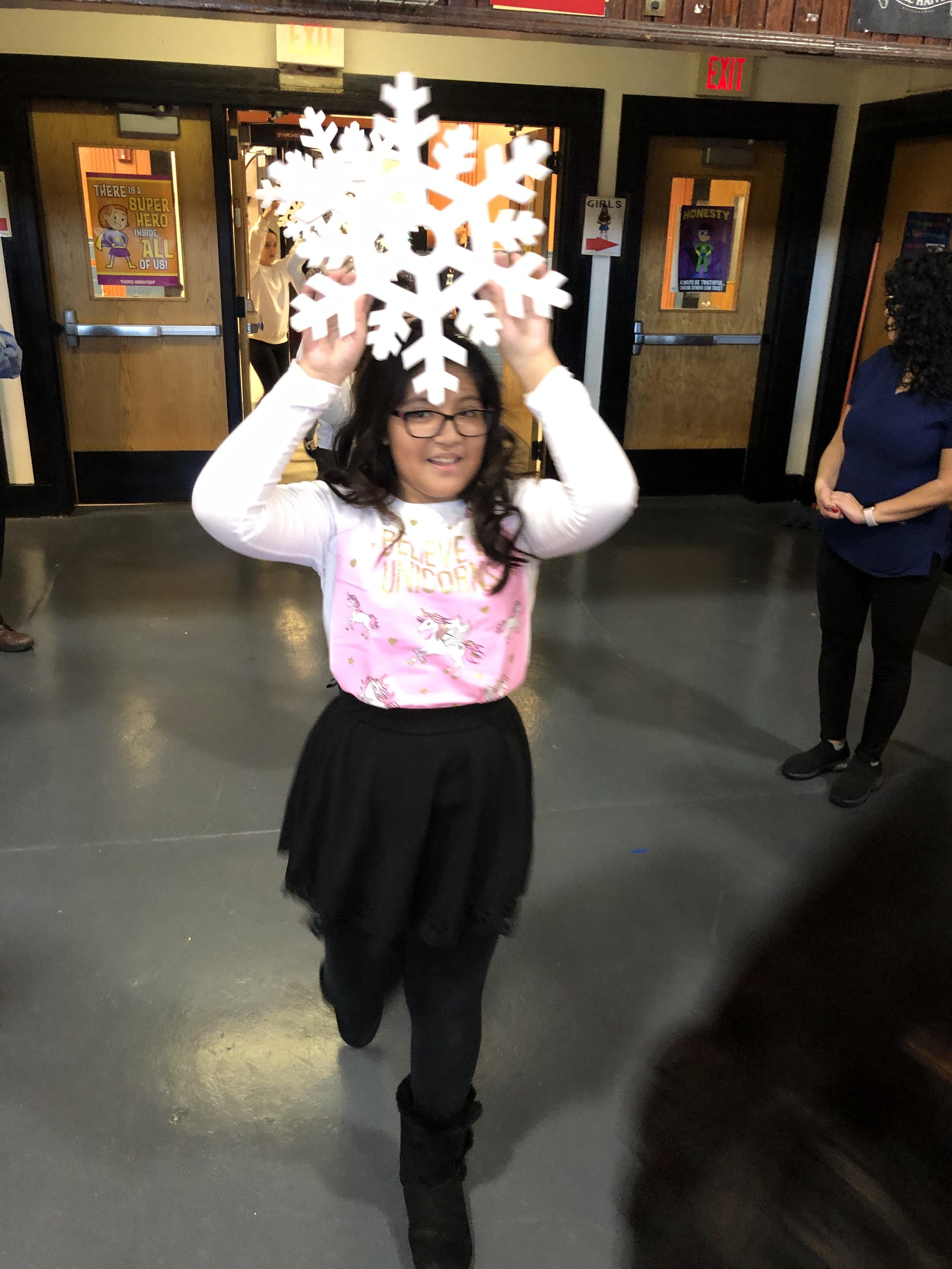 girl dressed with a pink top black leggings holding a white paper snowflake above her head entering the gym
